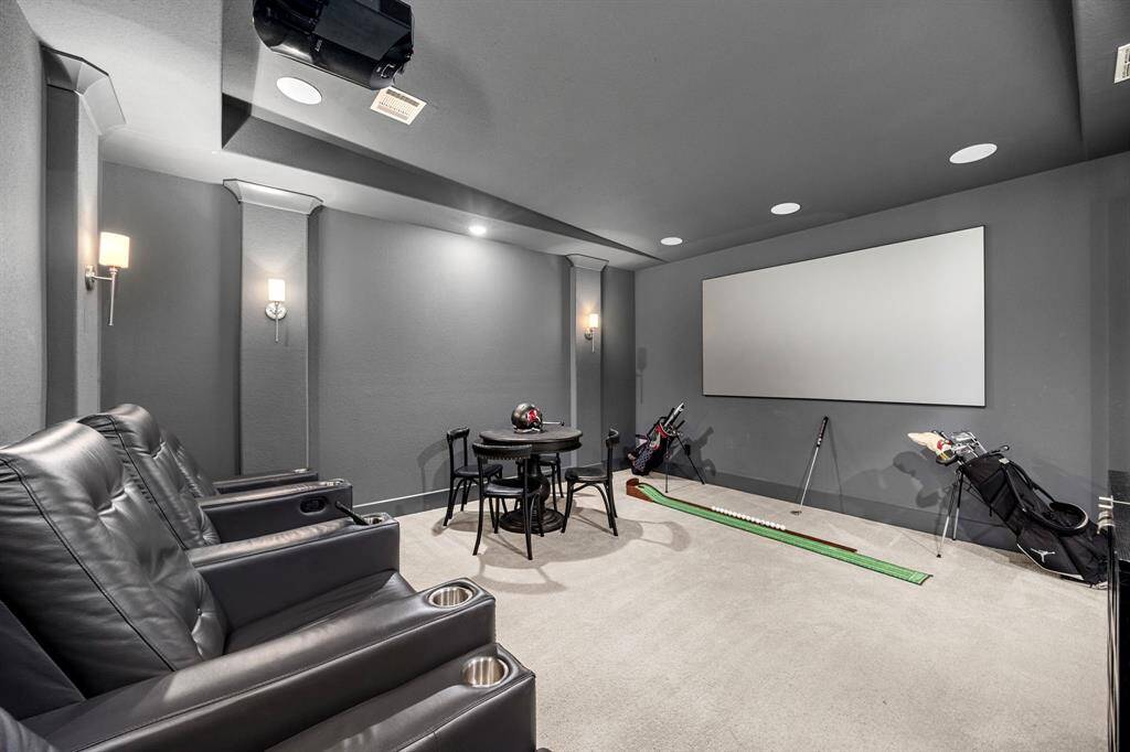 This photo showcases a home theater room with comfortable leather seating, a large screen, and a small table. The ambiance is cozy with wall sconces, and there's space for a golf putting practice setup, making it a versatile entertainment area. Projector is excluded