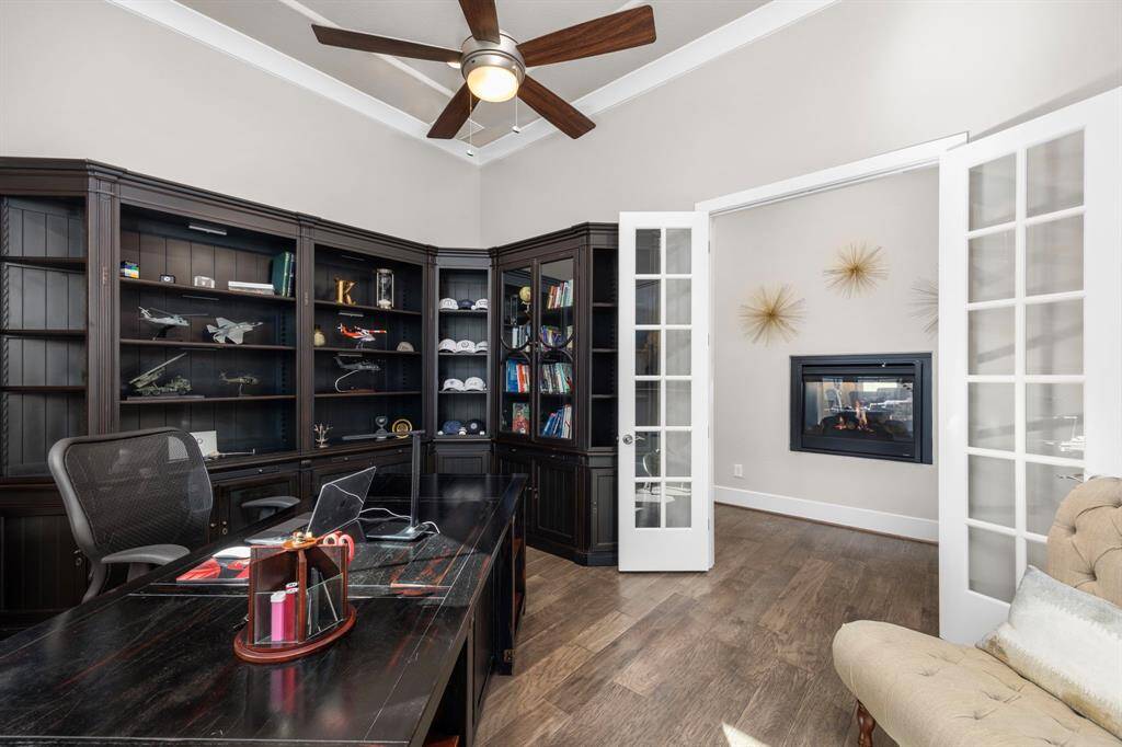 This photo showcases a well-appointed home office with dark wood built-in bookshelves and a desk. It features French doors, a cozy fireplace, and a ceiling fan, creating a warm and inviting workspace.