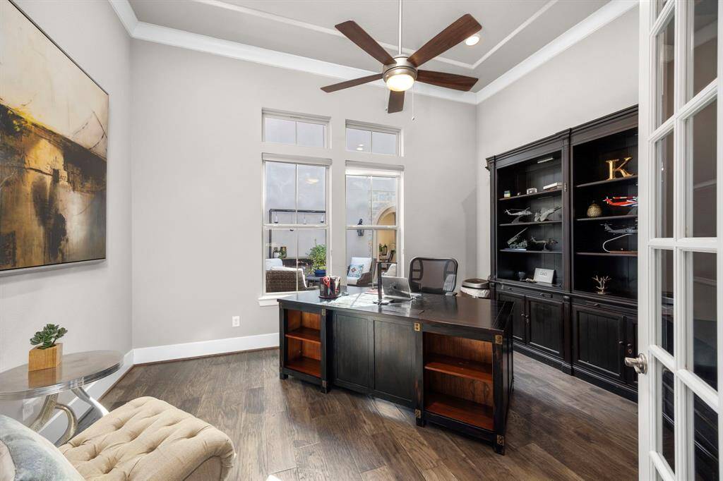 This photo showcases a bright home office with high ceilings and a ceiling fan. It features a large window, a sleek dark wood desk, and matching built-in shelving. The room is elegantly decorated with art and a simple plant, providing a professional and inviting workspace.