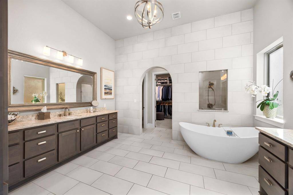 This spacious bathroom features a modern freestanding tub, dual sinks with granite countertops, and ample cabinetry. An archway leads to a walk-in closet, and a large window brings in natural light, complementing the elegant tile flooring.