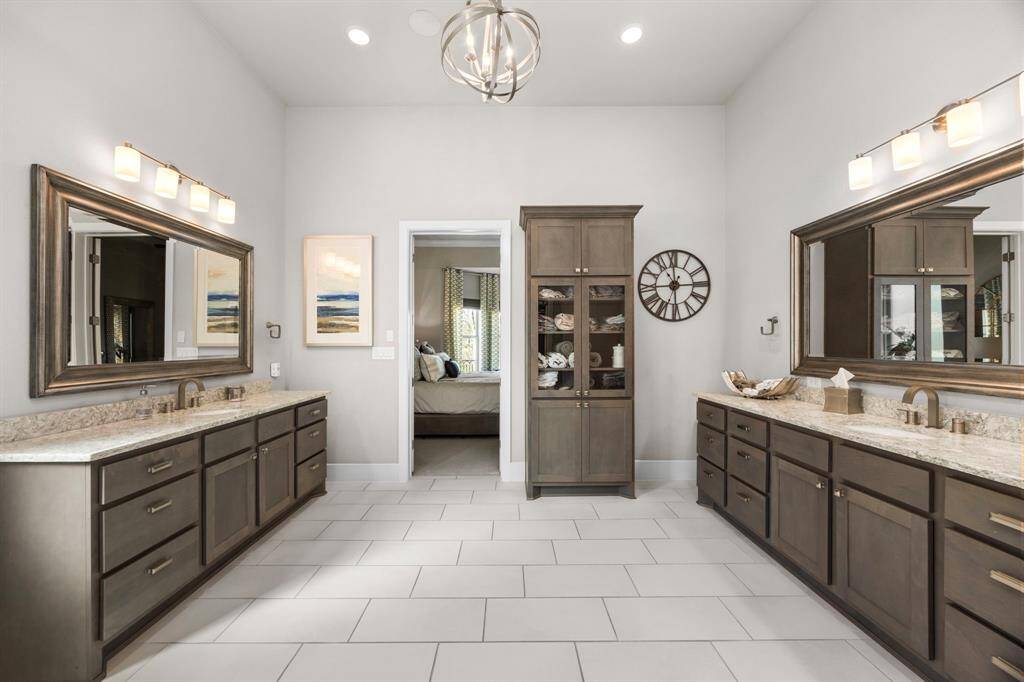 This spacious bathroom features dual vanities with granite countertops, elegant lighting fixtures, and ample cabinet storage. The neutral tile floor and modern finishes create a luxurious and functional space, with a doorway leading to a bedroom.