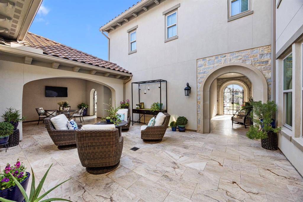 This inviting courtyard features elegant Travertine flooring, cozy wicker seating, and lush greenery, perfect for outdoor relaxation. A covered area with a mounted TV and arched entryways adds charm and functionality.