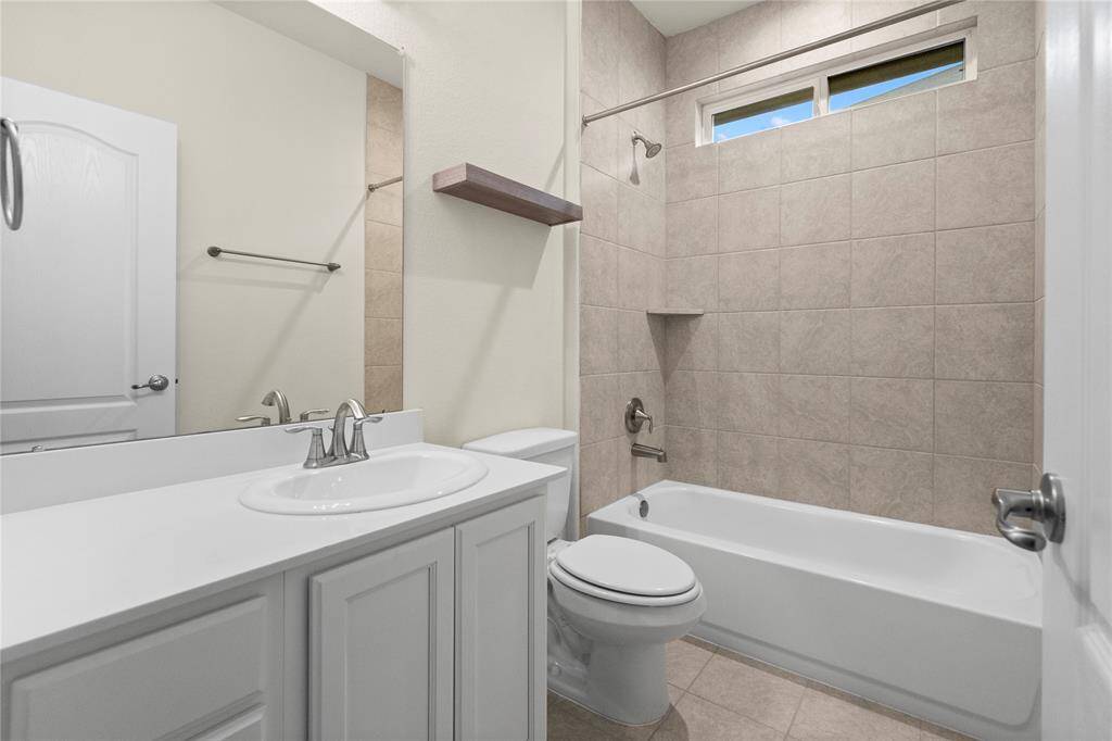 This secondary bathroom features a sleek design with a white vanity, a polished sink, and a full bathtub with a tiled shower area. It includes a small window for natural light, a mounted shelf for storage, and a neutral color palette for a clean and inviting look.