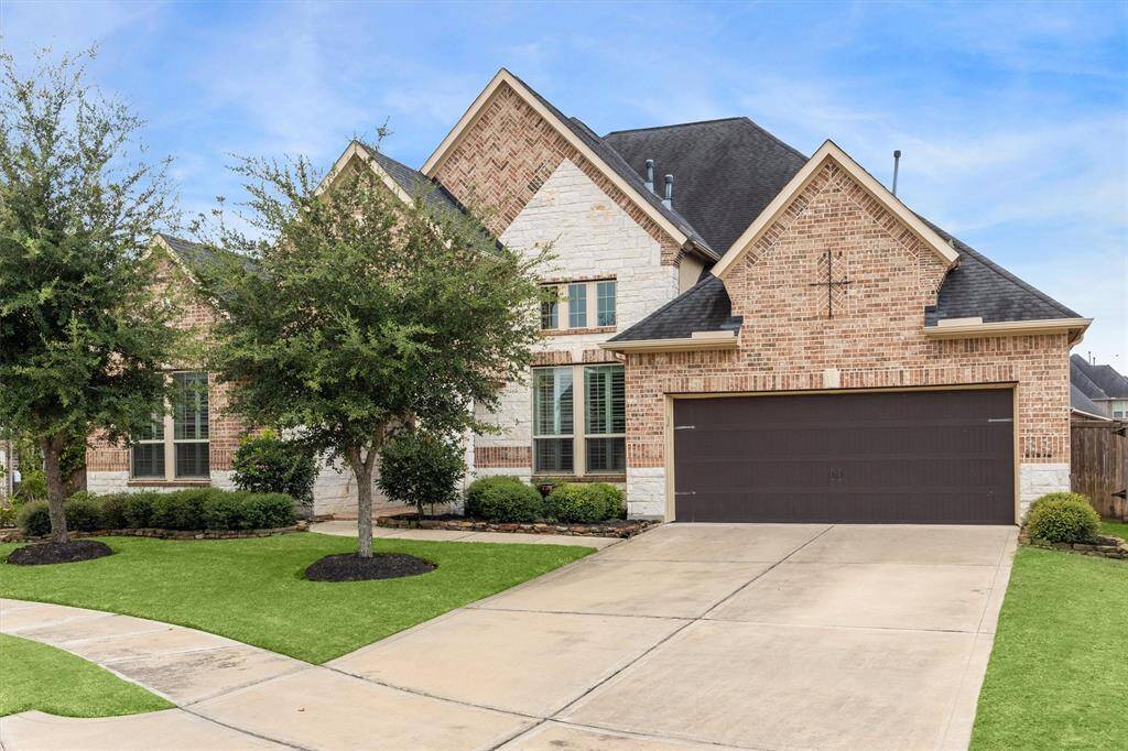 This is a single story brick home featuring a well-manicured lawn, mature trees, and an attached two-car garage. The home's architecture includes multiple gabled roofs and large windows.