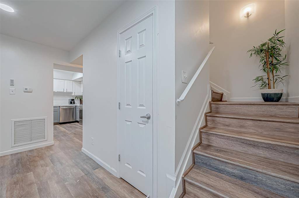 This photo shows a modern, well-lit entryway with wood flooring, a staircase with a sleek railing, and access to a kitchen featuring stainless steel appliances and white cabinetry. A potted plant adds a touch of greenery.