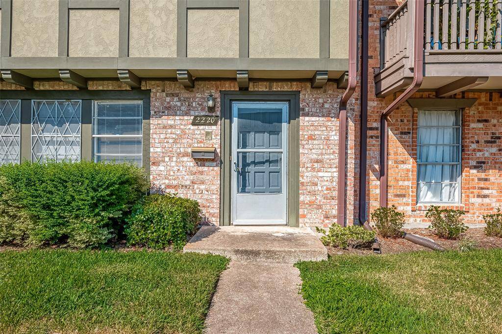 This photo shows the front entrance of a townhouse with a charming brick exterior. It features a well-maintained lawn, shrubs, and a blue front door with an address plaque. The design includes decorative window grilles and a small balcony above.