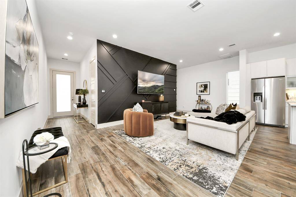 This modern living room features sleek wood flooring, a stylish black accent wall with a mounted TV, and ample natural light. The open layout includes a cozy seating area with a contemporary sofa and chairs, and a spacious kitchen with stainless steel appliances. A large rug and chic decor complete the inviting ambiance.