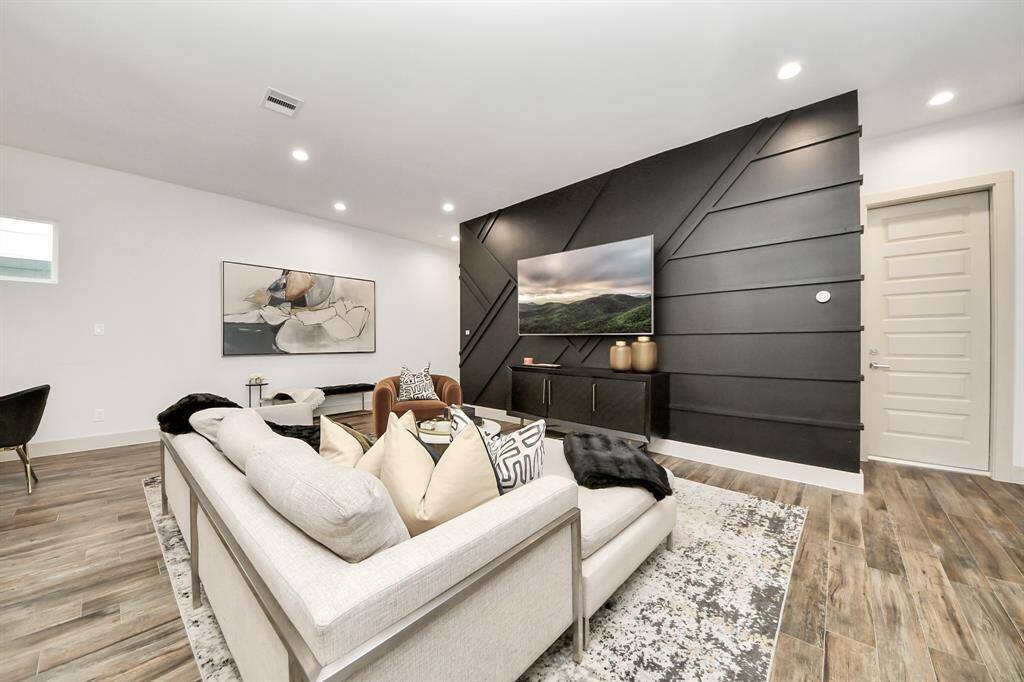 This modern living room features a sleek black accent wall with a mounted TV, stylish wood flooring, and a cozy sectional sofa adorned with decorative pillows. Recessed lighting and contemporary artwork enhance the inviting atmosphere.