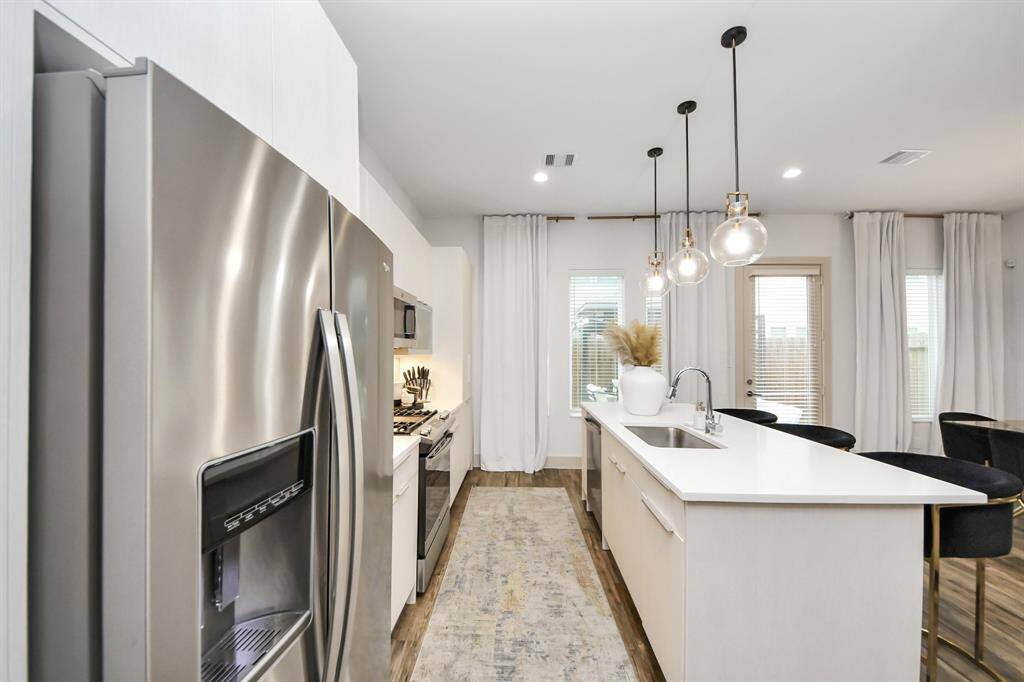This kitchen features a modern design with stainless steel appliances, a sleek island with a sink, and stylish pendant lighting. The space is bright and airy, with large windows and a door leading outside, complemented by elegant curtains.