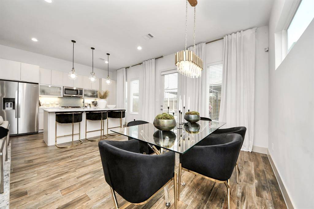 This modern open-concept kitchen and dining area features sleek stainless steel appliances, a spacious island with bar seating, elegant pendant lighting, and a chic glass dining table with plush black chairs. The space is bright and airy, accentuated by large windows and light wood flooring.