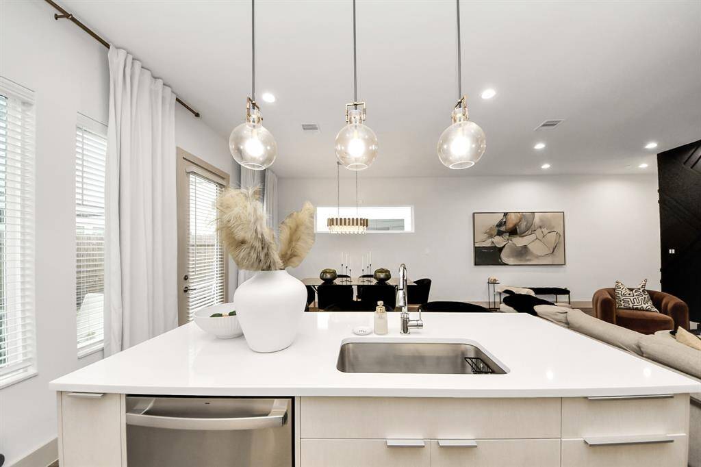 This modern kitchen features a sleek, open design with a large white island, pendant lighting, and integrated sink. Adjacent is a cozy living area with stylish decor and a neutral color palette, creating a bright and inviting atmosphere.