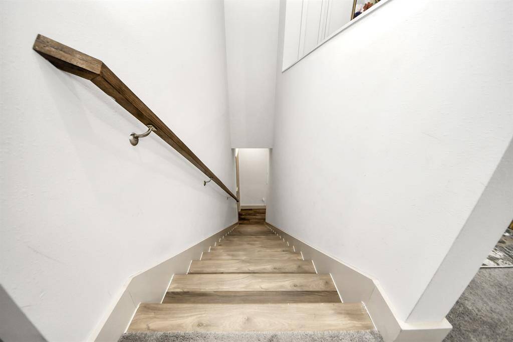 This photo shows a narrow, well-lit staircase with light wood steps and a sleek wooden handrail on a white wall, leading down to another level.
