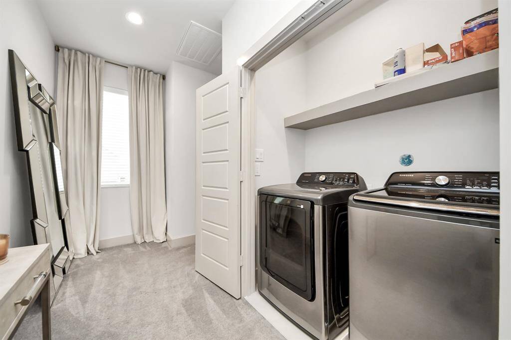 This photo showcases a modern laundry room with stainless steel washer and dryer. The space features a shelf for storage, a window with curtains allowing natural light, and a sleek mirror leaning against the wall, all set on a neutral-toned carpet.