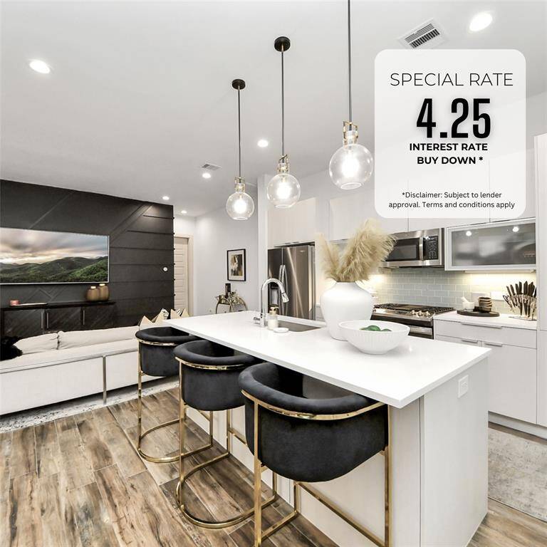 This modern open-concept space features a sleek kitchen with white cabinetry, a center island with stylish bar stools, and pendant lighting. The living area includes a cozy sofa and a wall-mounted TV, set against a chic, dark accent wall. Wood flooring and contemporary design elements provide a warm and inviting atmosphere.