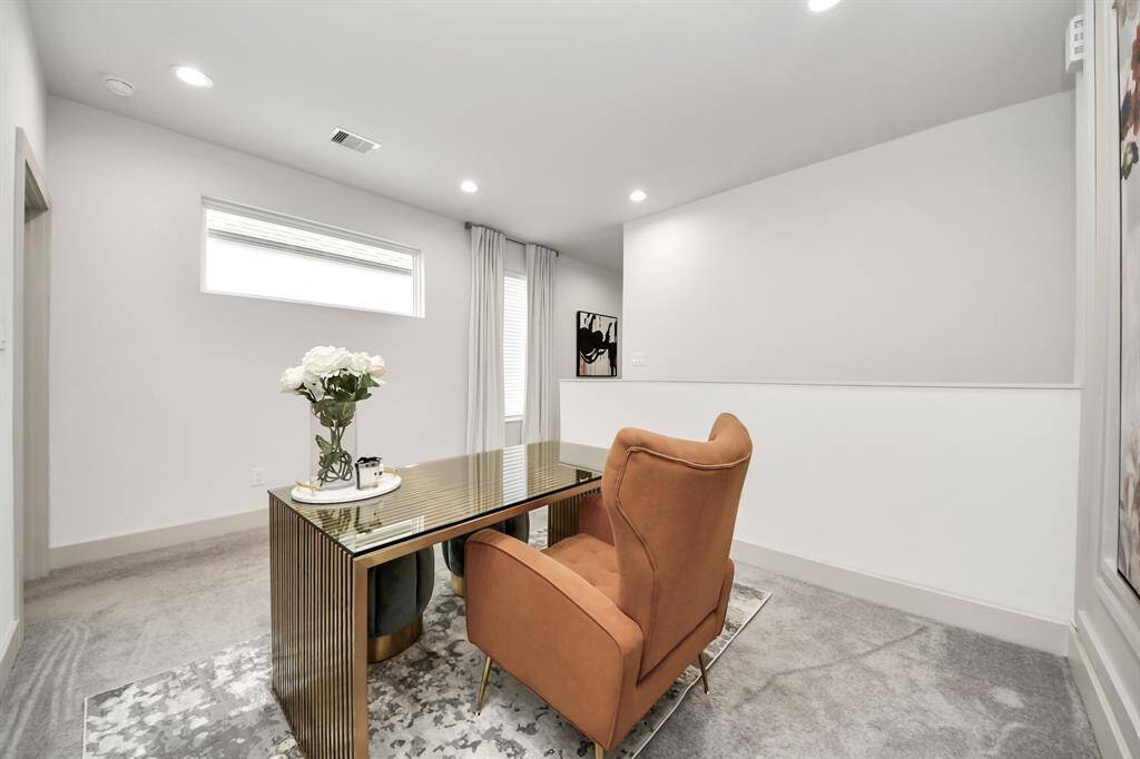 Bright home office with a modern design, featuring a sleek desk, comfortable chair, and soft carpeting. The room is well-lit with recessed lights and natural light from a high window.
