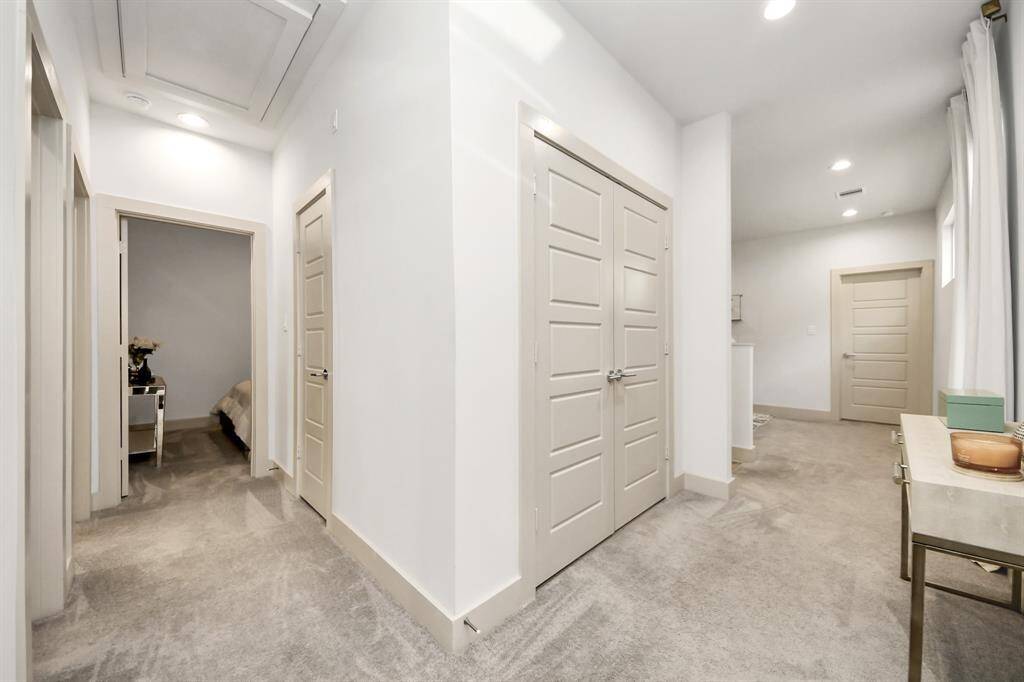 This photo showcases a modern, well-lit hallway with neutral tones and carpeted flooring. It features multiple doors leading to different rooms, a small table with decorative items, and recessed lighting, creating a clean and inviting atmosphere.