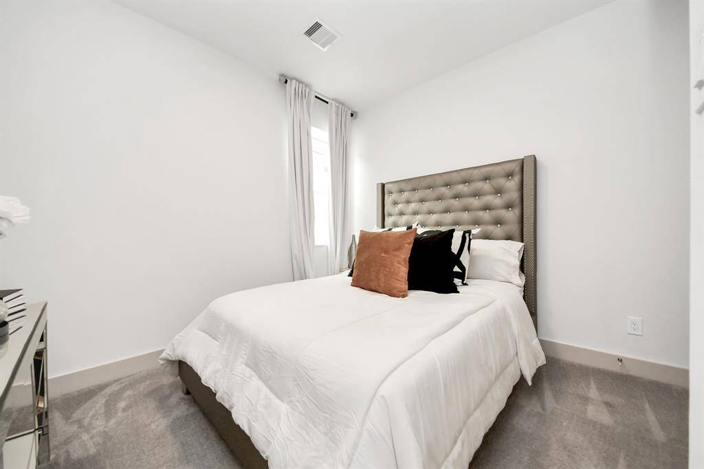 This cozy bedroom features a plush bed with an elegant tufted headboard, neutral walls, and ample natural light from a window with white curtains. A mirrored side table adds a touch of sophistication.