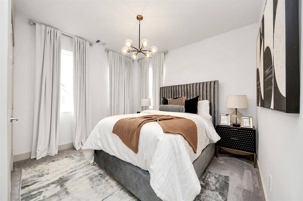 This bedroom features a modern design with a plush gray headboard and elegant bedding. Large windows with light curtains provide natural light, while a stylish overhead light fixture and contemporary art add sophistication. A cozy rug and bedside tables with lamps complete the inviting space.