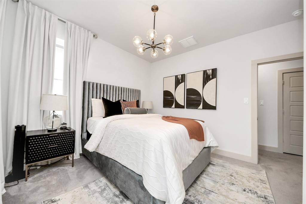 This bedroom features a modern design with a plush gray bed, stylish geometric artwork, and a chic light fixture. The space is bright and airy, with white walls and a large window dressed in curtains, complemented by a sleek bedside table with a lamp.