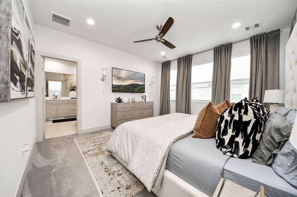 This spacious bedroom features a modern design with neutral tones, a comfortable bed, and large windows for natural light. It includes a ceiling fan, wall-mounted TV, and stylish decor. The room connects to a sleek en-suite bathroom, enhancing convenience and privacy.