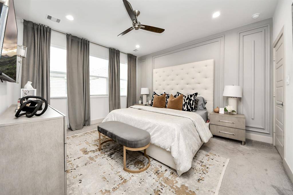 This bedroom is elegantly designed with a neutral color palette, featuring a large bed with a tufted headboard, modern side tables with lamps, and a stylish bench at the foot. Natural light streams through three windows, accented by floor-length curtains. The room also includes a ceiling fan and cozy carpeting, creating a comfortable and inviting atmosphere.