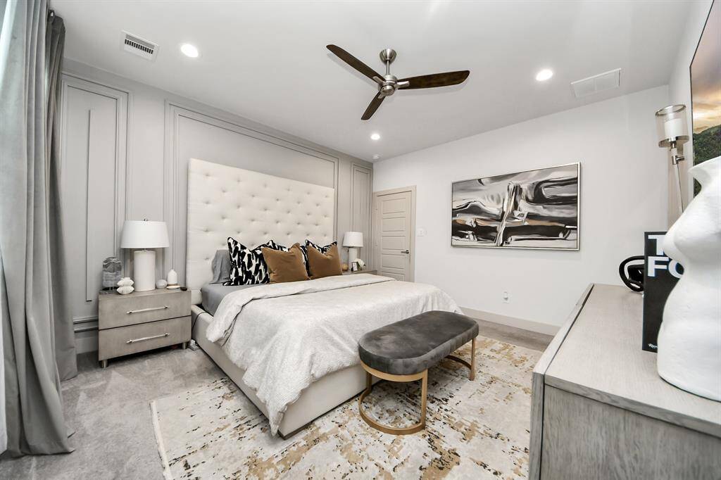 This bedroom features a modern, elegant design with a plush bed, stylish artwork, and a neutral color palette. It includes a ceiling fan, soft lighting, and ample space, creating a comfortable and inviting atmosphere.