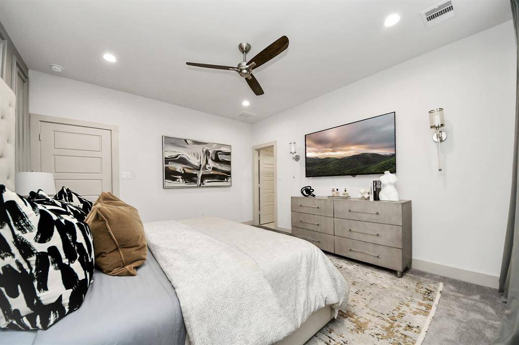 This bedroom features a modern design with neutral tones. It includes a cozy bed with decorative pillows, a stylish ceiling fan, and ample lighting. A large dresser provides storage, and the space is accented with contemporary artwork and a wall-mounted TV.