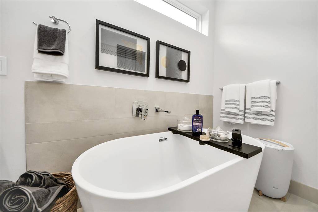 This bathroom features a sleek, modern design with a freestanding bathtub, neutral-toned tiles, and minimalistic decor. It offers ample natural light and stylish accents like framed artwork and neatly arranged towels, creating a relaxing, spa-like atmosphere.