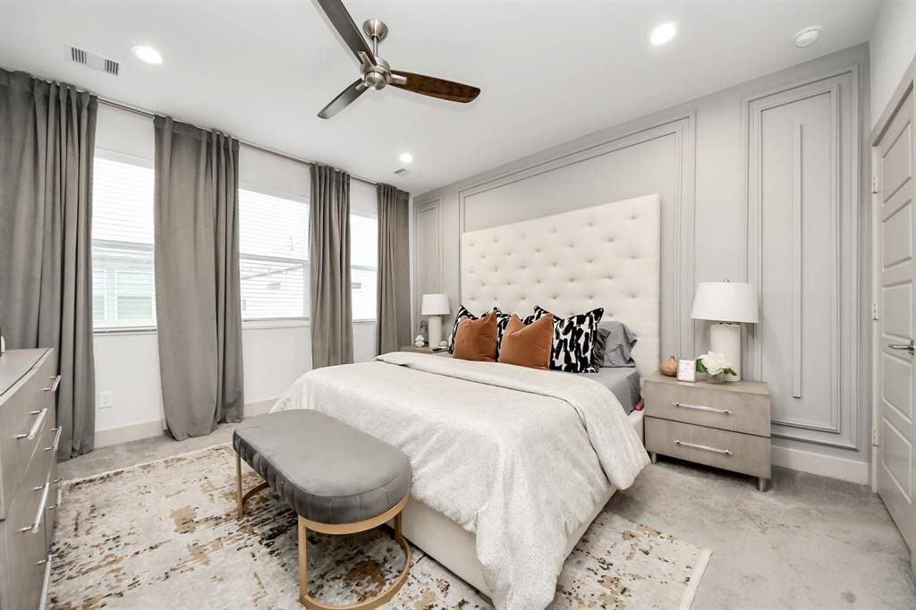 This bedroom features a modern design with a plush, tufted headboard and neutral tones. Large windows with elegant drapes provide ample natural light, and a ceiling fan adds comfort. The room includes stylish bedside tables and a bench at the foot of the bed, creating a cozy yet sophisticated atmosphere.