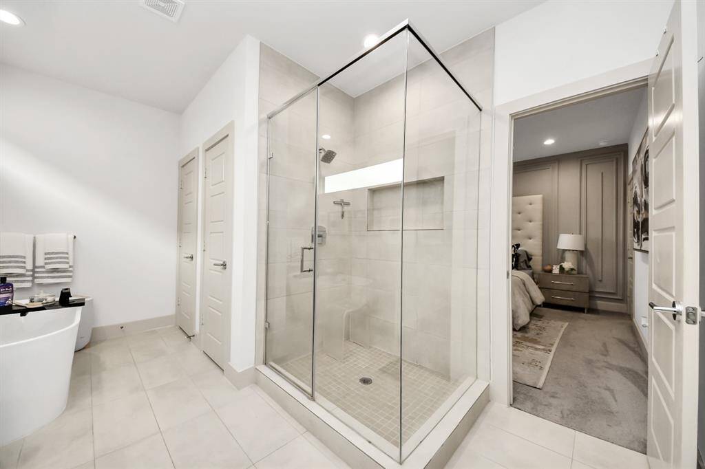 This photo showcases a modern bathroom featuring a spacious glass-enclosed shower, a sleek freestanding tub, and neutral tile flooring. There's an adjacent bedroom visible through an open door, adding to the seamless layout.