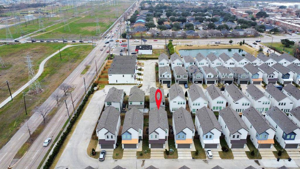 This aerial photo shows a modern housing development with rows of similar-styled homes. The homes have neatly arranged driveways, and there's a marker indicating a specific property. The neighborhood is adjacent to a major road and open green space, with a pond visible nearby.