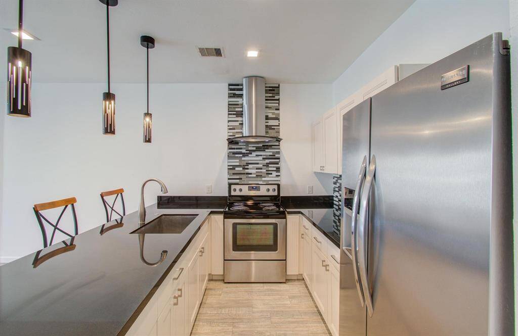 Modern kitchen with stainless steel appliances, sleek black countertops, and stylish pendant lighting. Features a contemporary backsplash and a spacious layout with an island bar seating.