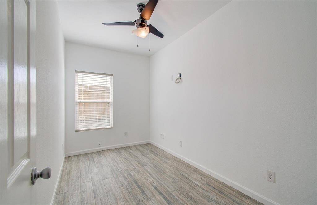 Bedroom features a clean, minimalist design with white walls and vinyl flooring. It has a ceiling fan and a window that allows natural light to brighten the space, making it ideal for an office or bedroom.