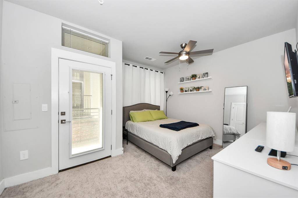This bedroom features a neutral color palette with plush carpeting. It includes a large bed, ceiling fan, and access to a small balcony through a glass door. The room is well-lit.