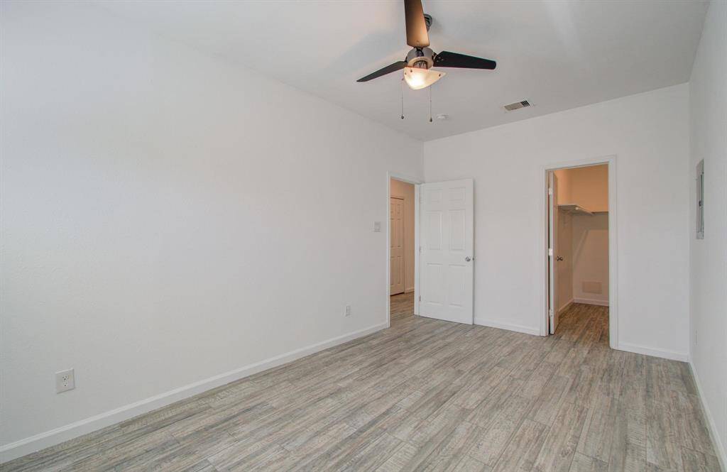 Another view of primary bedroom featuring a clean, modern design with vinyl flooring and white walls. It includes a ceiling fan for comfort and two doors leading to a hallway and a walk-in closet, offering ample storage space.