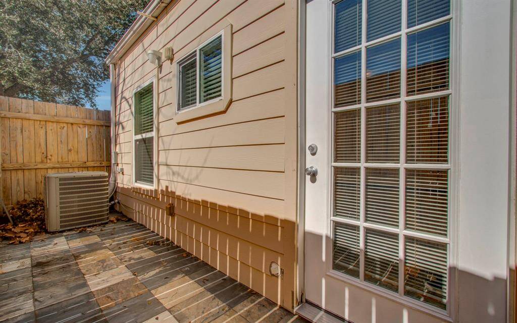 Small outdoor patio area with a tiled floor, a wooden fence for privacy, and a glass-paned door leading inside. It features two windows and a wall-mounted light fixture, providing a cozy space for relaxation.