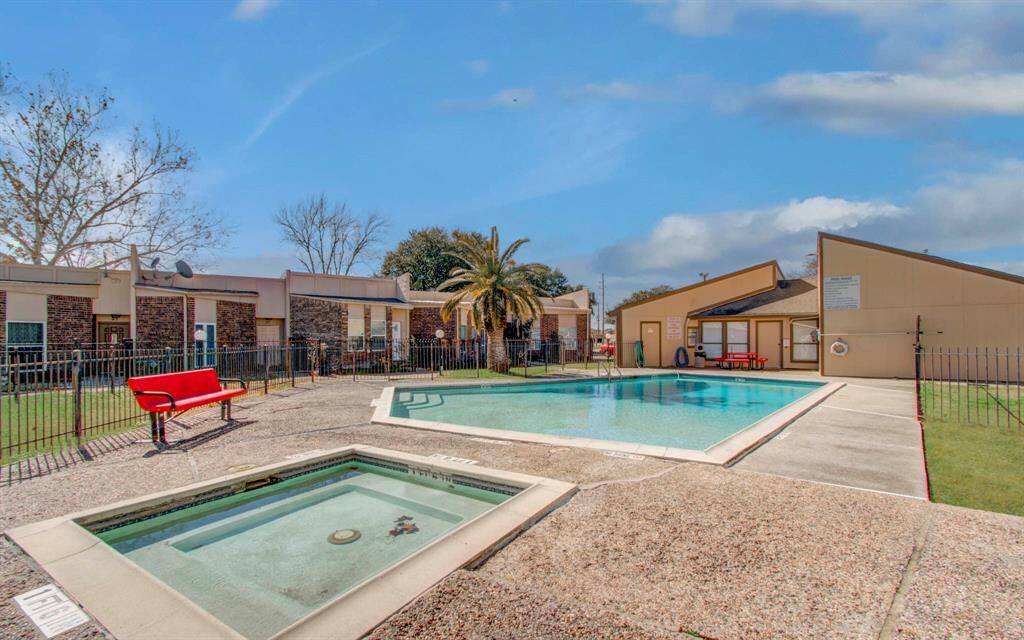 Charming community pool area featuring a spacious swimming pool, a hot tub, and a sunlit seating area with benches, surrounded by well-maintained landscaping.