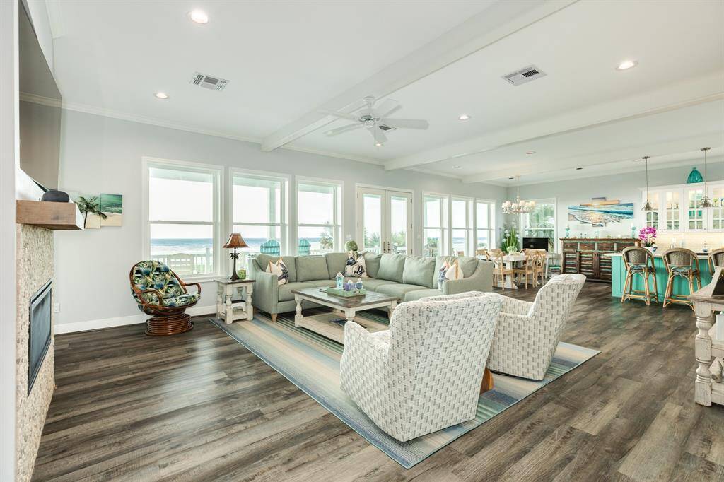 Interior view of living room/dining area & kitchen island. Home features 10' ceilings, exposed beams & LVP flooring throughout. Wall of windows and french doors allow for an abundance of natural light & provide wonderful views dunes & beach.