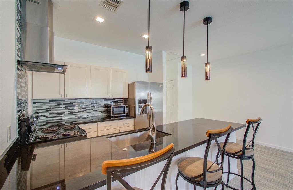 Modern kitchen features sleek black countertops, stylish tile backsplash, and elegant pendant lighting. Includes a stainless-steel refrigerator, an electric stove, and a convenient breakfast bar with seating for three. The bright, open space is perfect for entertaining and everyday cooking.