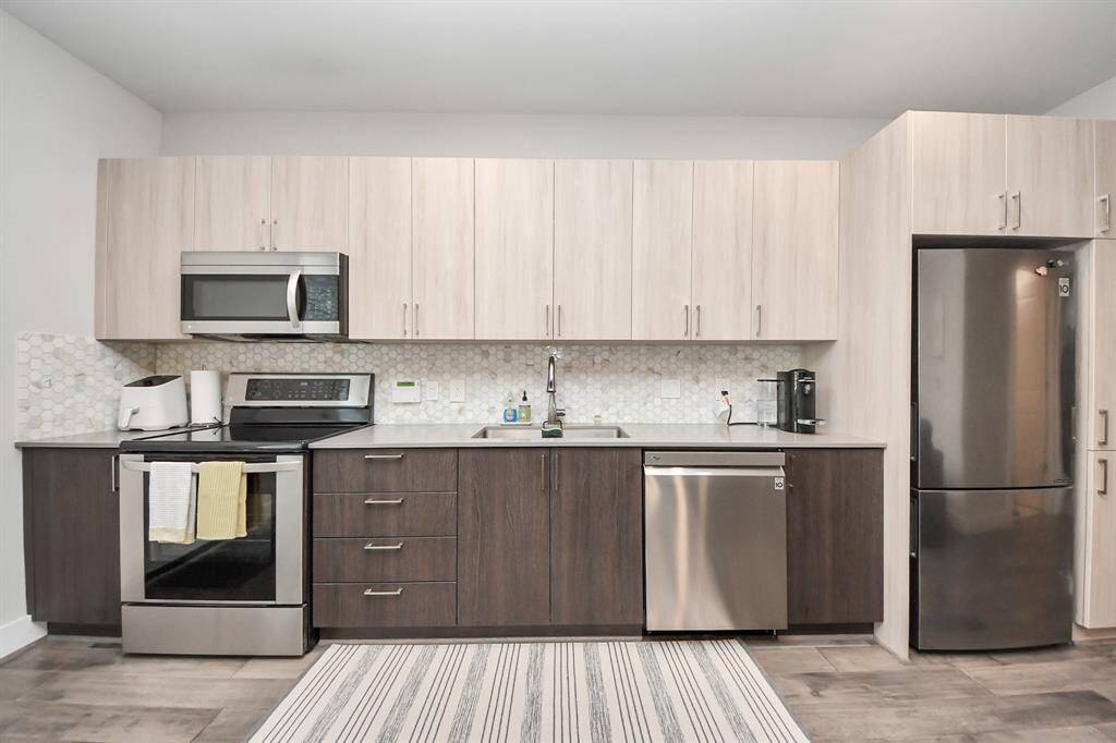 Modern kitchen with sleek stainless steel appliances, light wood cabinets, and a hexagonal tile backsplash. It features a built-in microwave, oven, and dishwasher.