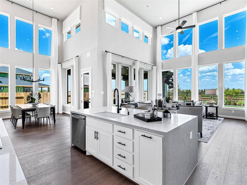 This photo showcases a modern, open-concept living space with high ceilings and expansive windows that fill the area with natural light. The kitchen features a large island with white cabinetry, while the dining area and living room offer a seamless flow, ideal for entertaining. The decor is sleek and contemporary, complemented by the bright, airy ambiance.