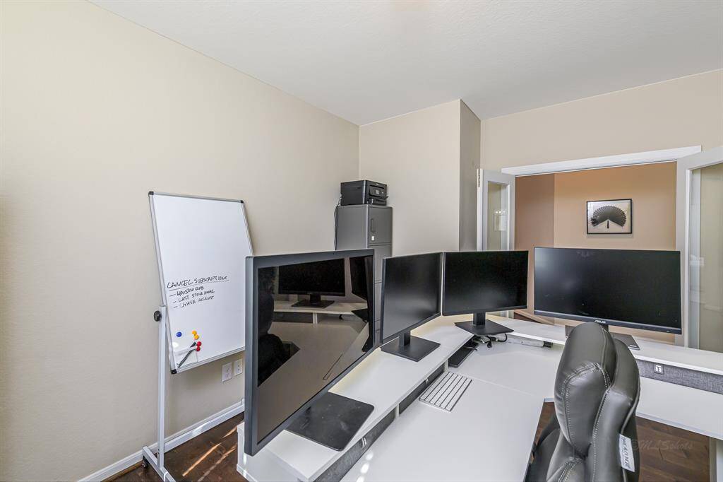 This room is set up as a home office with multiple monitors on a desk, a whiteboard, and a comfortable office chair. The space is bright with neutral walls and a practical layout, ideal for remote work or study.