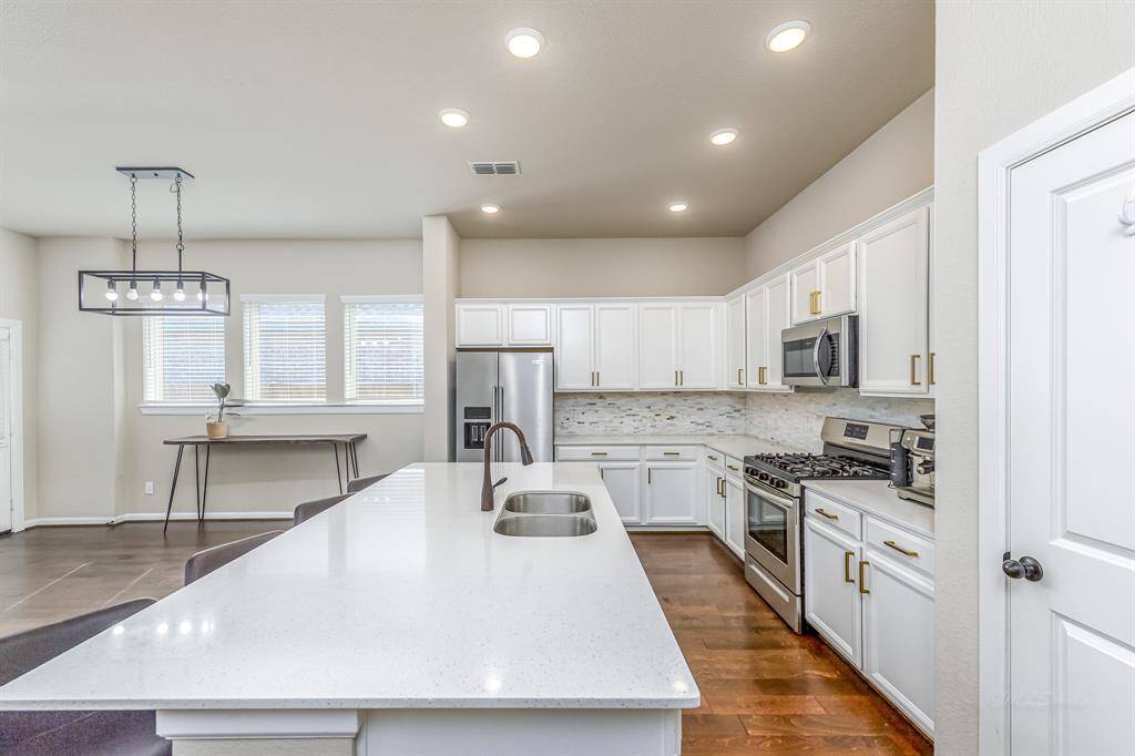 Modern kitchen with a spacious layout, featuring white cabinetry, stainless steel appliances, and a large island with a sink. The room is well-lit with recessed lighting and a stylish pendant fixture.