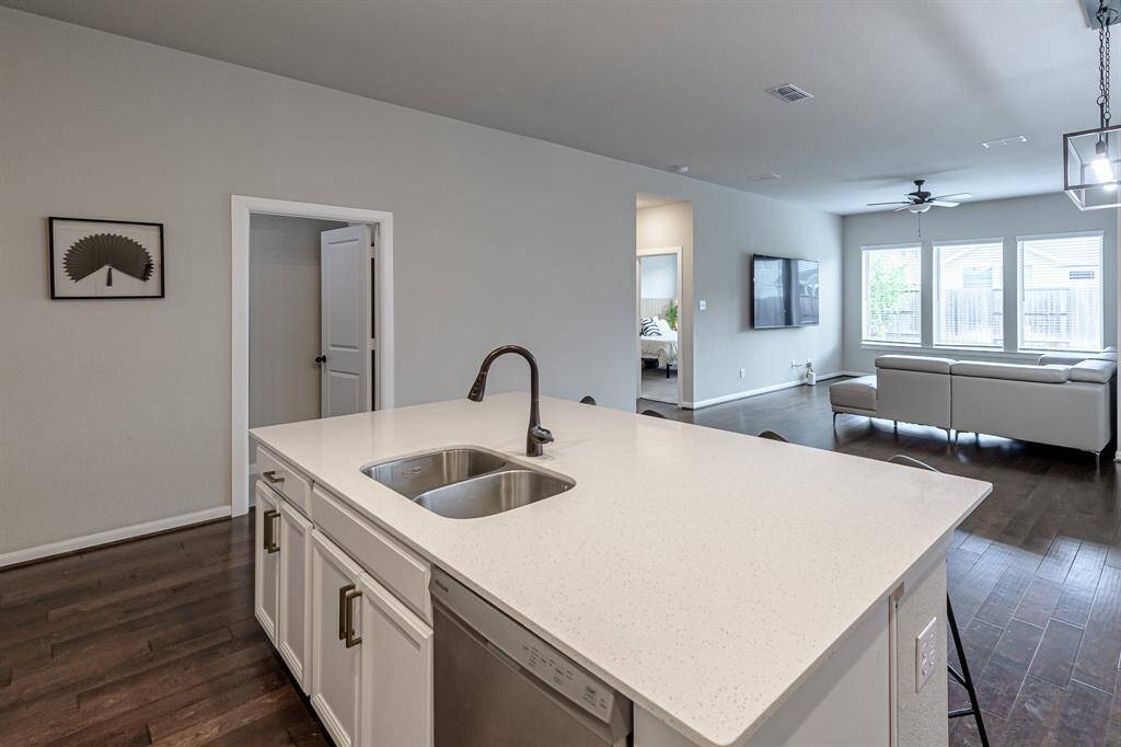 Modern open-concept living space featuring a spacious kitchen with a large island, light countertops, and a sink. The living area includes a comfortable sofa, large windows for natural light, and a ceiling fan. Dark wood flooring adds elegance.