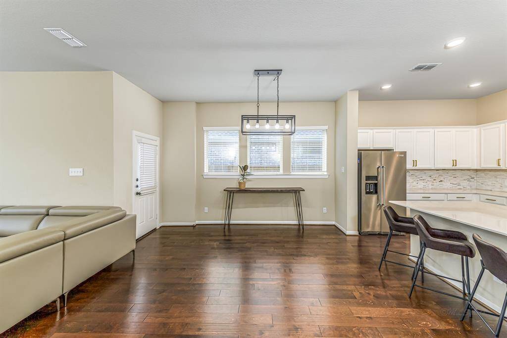 This open-concept living space features rich hardwood floors, a modern kitchen with stainless steel appliances, and a spacious island with seating. The room is bright and airy, with ample natural light and contemporary lighting fixtures.