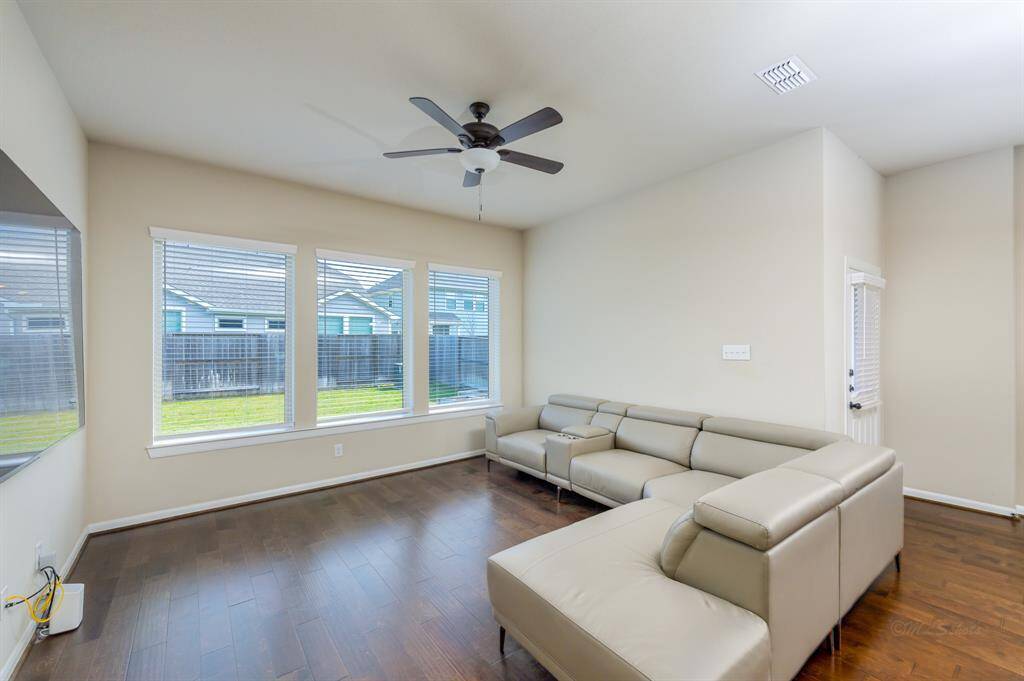 Bright living room with large windows, neutral walls, and dark wood flooring. Features a modern ceiling fan and a spacious sectional sofa. Ideal for relaxation and entertaining.