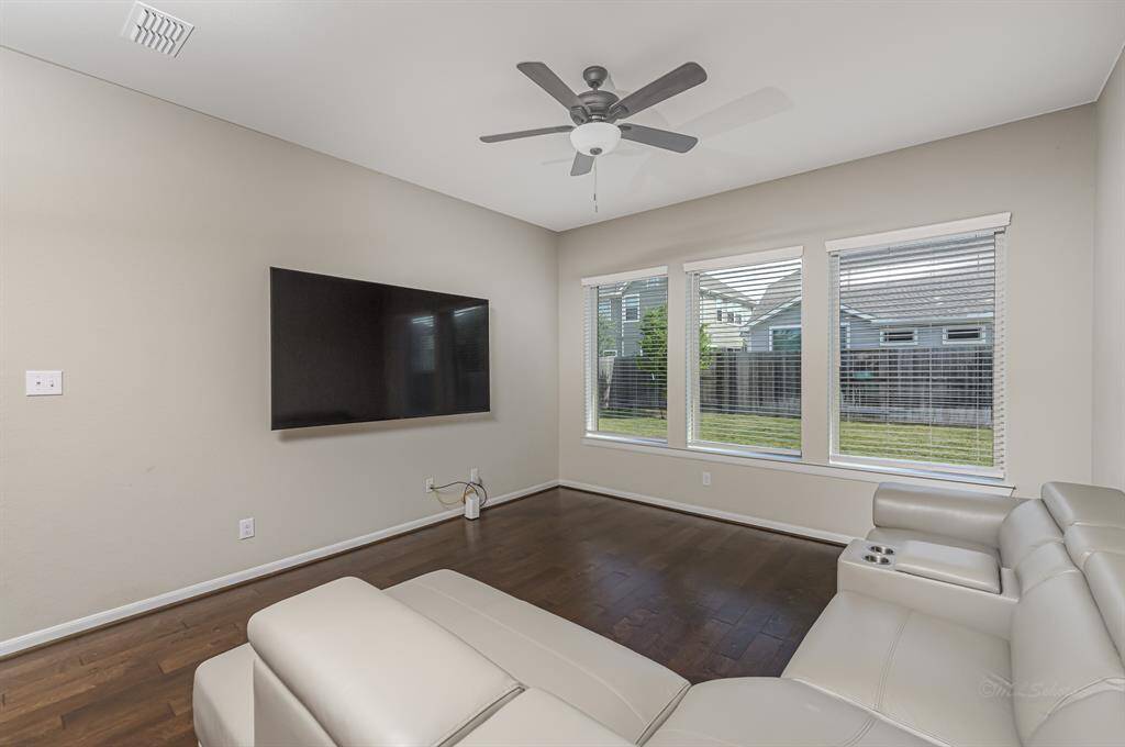 This living room features hardwood floors, a wall-mounted TV, and a ceiling fan. Large windows provide ample natural light and offer views of the backyard.