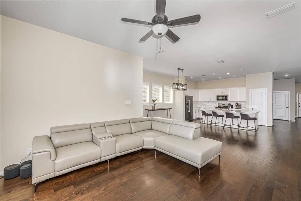 This open-concept living area features a spacious layout with a modern kitchen, complete with a large island, stainless steel appliances, and white cabinetry. The living room has a sleek sectional sofa and ceiling fan, set on elegant hardwood floors. Natural light fills the space, creating a welcoming atmosphere.