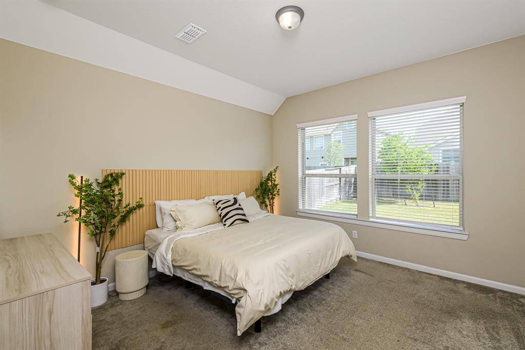 This bedroom features neutral tones, a cozy bed with a stylish headboard, and ample natural light from two large windows overlooking a backyard. The space is accented with potted plants and a minimalist dresser, creating a peaceful retreat.