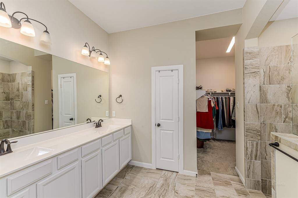 Spacious bathroom with dual sinks, elegant lighting, and a large mirror. Features a walk-in closet and a beautifully tiled shower area.